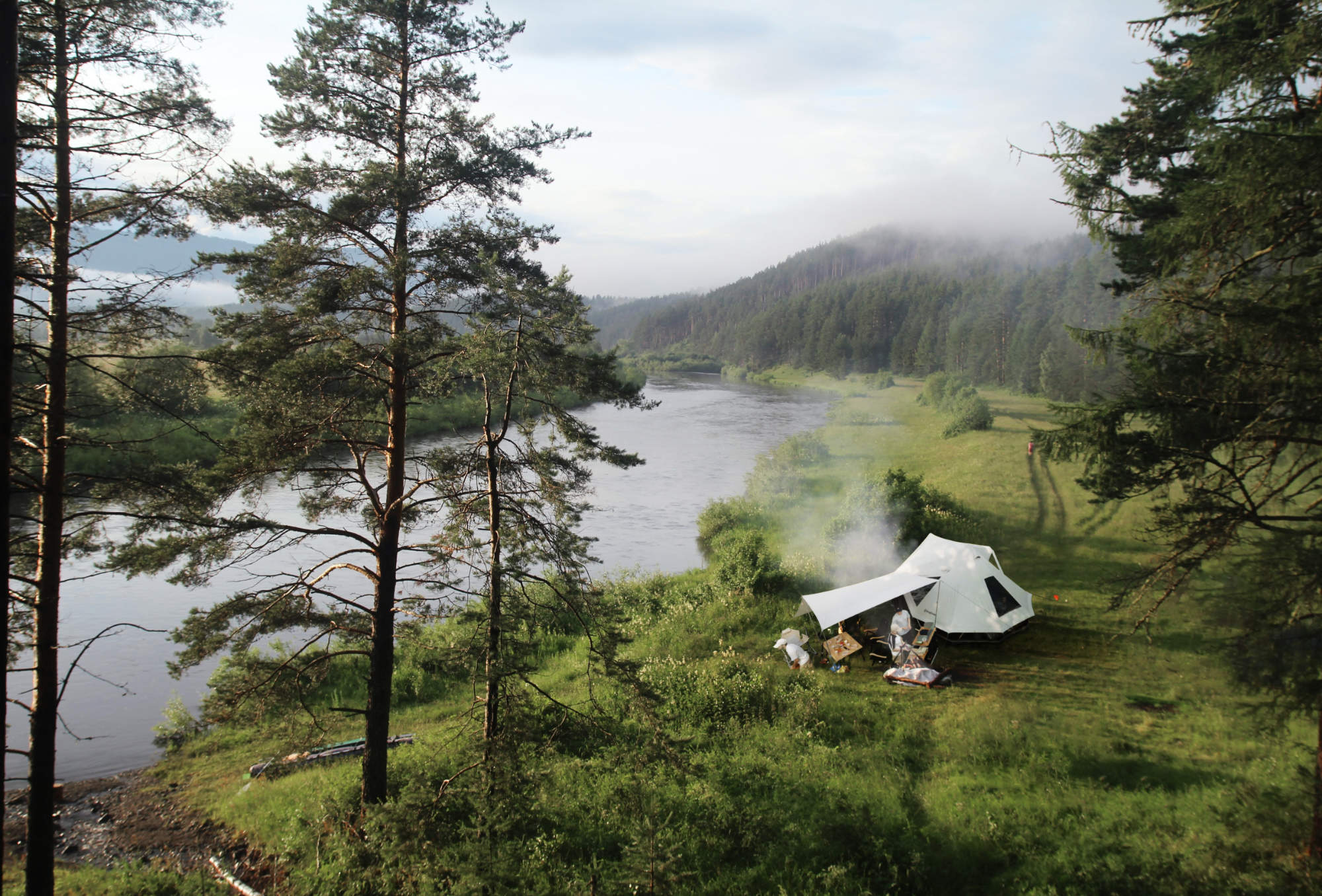 Zeltlager mit Rauchentwicklung am Flussufer in einer bewaldeten Berglandschaft, aufgenommen aus erhöhter Perspektive. Zu sehen ist das Baumwollzelt Khan Villa.
