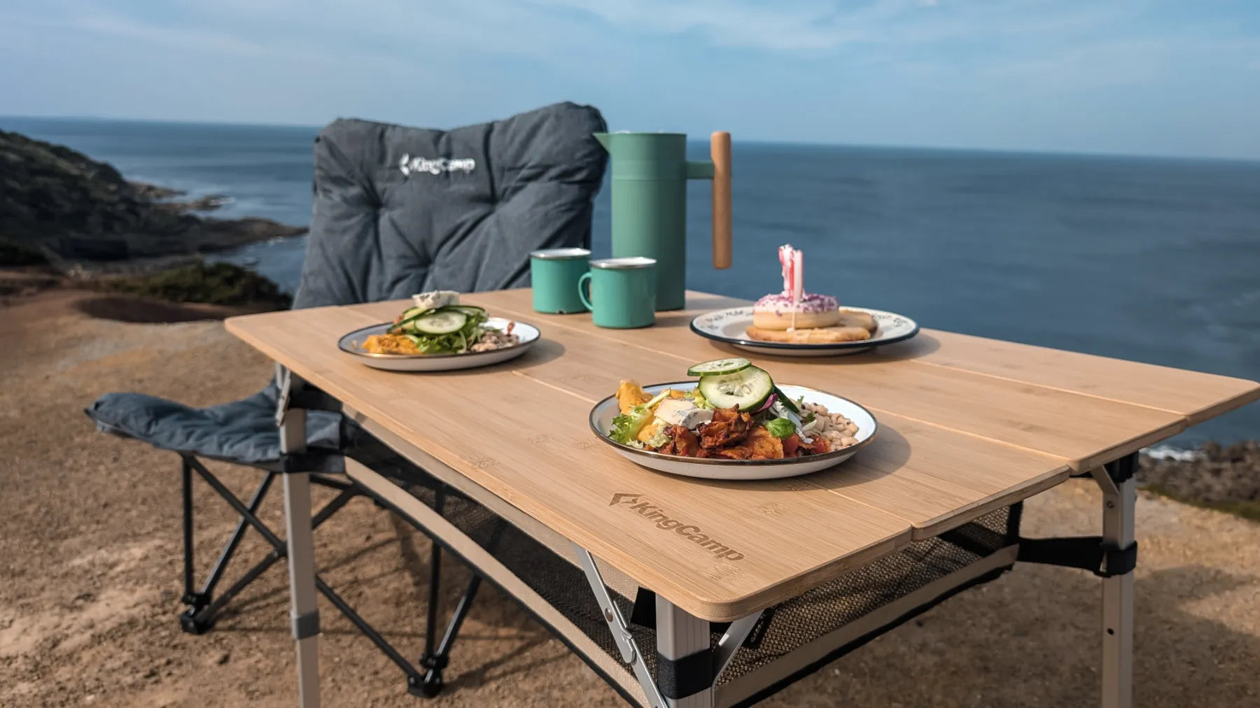 Ein KingCamp-Tisch mit gedeckten Tellern und Emaillebechern steht vor einem Campingstuhl mit Blick aufs Meer.