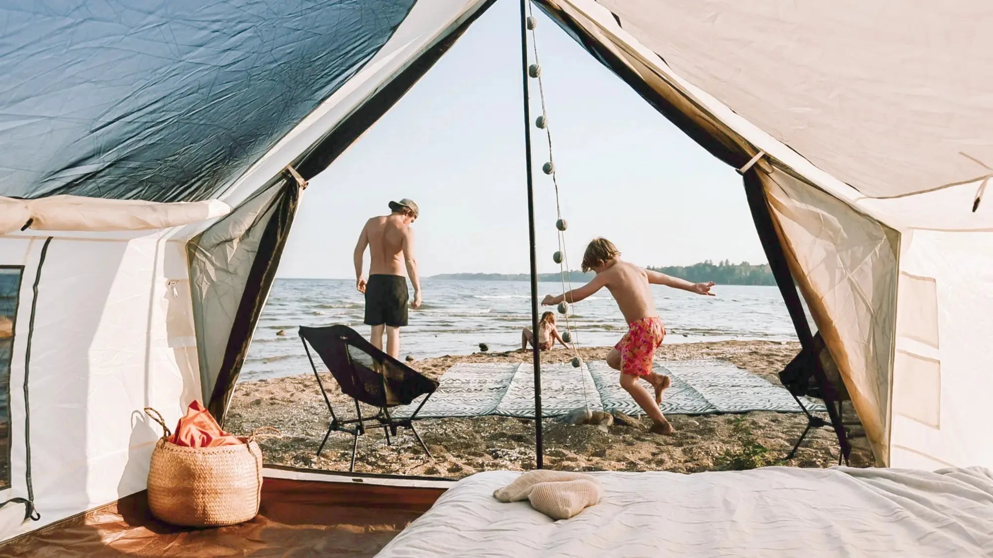 Blick aus einem offenen Zelt auf einen Strand, an dem ein Kind spielt, ein Mann steht und ein weiteres Kind im Sand sitzt.